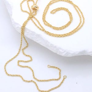 Puede incluir: Un collar de cadena en tono dorado con una delicada textura de cuerda trenzada. La cadena incluye un pequeño componente deslizante redondo y liso para ajustar la longitud, con dos extremos abiertos terminados en pequeños anillos circulares.