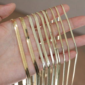Peut inclure: Une collection de plusieurs chaînes à chevrons dorées présentées en différentes largeurs. Chaque chaîne présente un design à maillons plats et imbriqués qui crée une surface lisse et réfléchissante. Les chaînes sont disposées côte à côte.