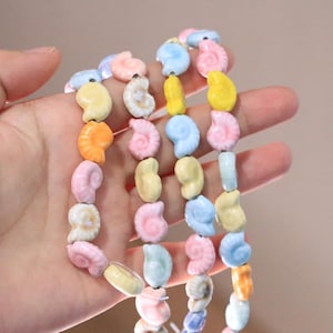 Könnte beinhalten: Eine Sammlung kleiner, spiralförmiger Muschelperlen in Pastellfarben wie Hellblau, zartem Rosa, Blassgelb und Orange. Diese keramikartigen Perlen sind auf einen dünnen weißen Faden aufgezogen und weisen eine glatte, glänzende Oberfläche auf.