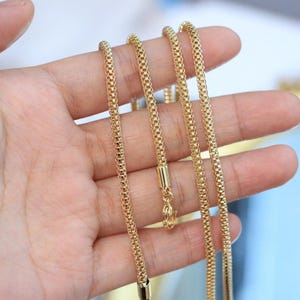 Könnte beinhalten: Eine goldfarbene Kettenhalskette mit einem abgerundeten, texturierten Box-Link-Design. Die Kette hat eine polierte Oberfläche und enthält eine zylindrische Endkappe mit einem kleinen Karabinerverschluss, die ein gleichmäßiges, quadratisches Muster zeigt.