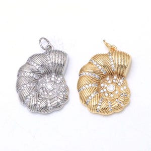 Könnte beinhalten: Zwei muschelförmige Anhänger, einer in Silberoptik und einer in Goldoptik, beide mit einer gerippten Texturoberfläche und besetzt mit kleinen klaren Kristallen im gesamten Design, jeweils mit einem kleinen runden Ring zur Befestigung oben.