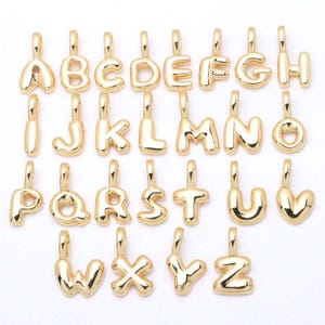 Pode incluir: Uma coleção de vinte e seis pingentes de letras em metal dourado, cada um com um design arredondado e volumoso e um pequeno laço no topo para fixação. O conjunto inclui o alfabeto completo de A a Z, organizado em quatro linhas distintas.