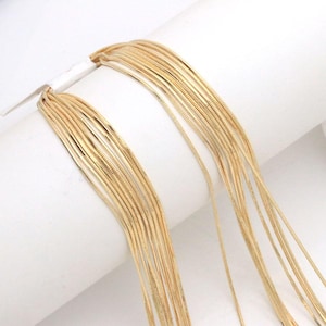 Könnte beinhalten: Eine mehrsträngige Schlangenkette in Goldoptik, bestehend aus zahlreichen dünnen, flexiblen Metallketten, die zusammenfallen. Die Ketten haben eine glatte, reflektierende Oberfläche und eine gleichmäßige, engmaschige Struktur für ein fließendes Aussehen.