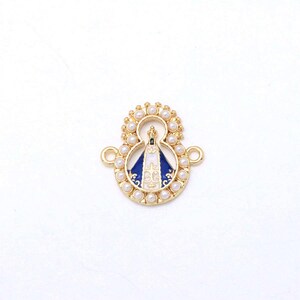 Pode incluir: Um pingente de metal dourado com a figura central de Nossa Senhora Aparecida com um manto de esmalte azul e branco, rodeado por uma auréola de pequenas pérolas brancas. O pingente inclui dois elos laterais para fixação em joias ou acessórios.