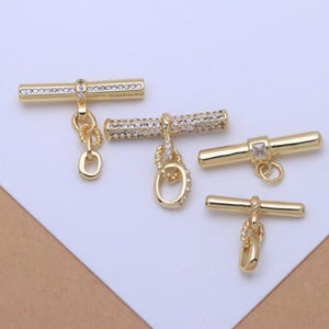May include: Four gold-toned toggle clasps for jewelry making. Two clasps feature cylindrical bars encrusted with clear stones and attached oval rings. One clasp has a smooth cylindrical bar with a single square stone, and one has a smooth bar.