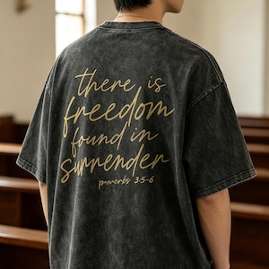 May include: A charcoal grey acid-wash t-shirt featuring gold cursive text on the back that reads, There is freedom found in surrender, followed by the smaller text, proverbs 3:5-6. The shirt has short sleeves and a crew neckline.