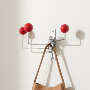 Op de afbeelding: Een wandkapstok met een verticale zilverkleurige metalen plaat met drie uitschuifbare chromen armen, elk bekroond met een gladde, glanzende rode bol. Een beige leren handtasriem hangt aan een van de chromen armen van de kapstok.