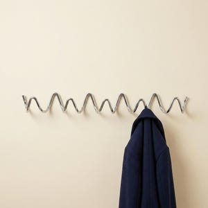 May include: A wall-mounted coat rack featuring a continuous, polished chrome metal wire bent into a repeating zigzag or wave pattern. A dark navy blue coat hangs from one of the lower curves of the metal structure, which is secured at both ends.