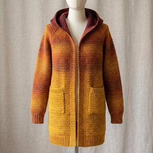 Könnte beinhalten: Eine langärmelige, vorne offene Häkelstrickjacke mit Kapuze und zwei Vordertaschen. Das Kleidungsstück zeigt ein Farbverlaufsmuster, das von einem tiefen Burgunderrot an der Kapuze zu einem leuchtenden Orange und einem hellen Gelb am Saum übergeht.