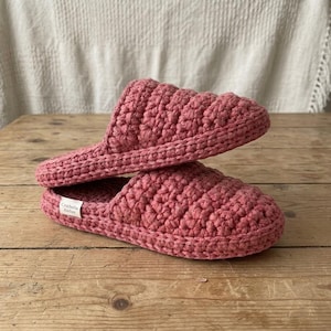 May include: A pair of dusty rose crochet slippers featuring a textured, thick yarn construction with a rounded toe design. A small white rectangular fabric label with the black text "Crochella Atelier" is attached to the side of the bottom slipper.