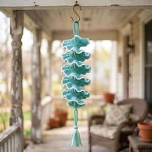May include: A hanging crochet decoration featuring a vertical series of ruffled, teal-coloured circular layers with white edges. The piece is suspended from a metal S-hook and terminates in a multi-coloured tassel at the bottom, showcasing intricate yarn work.