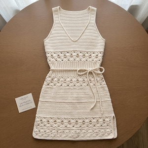 May include: A sleeveless cream-colored crochet dress featuring a V-neckline, a waist tie, and repeating floral patterns. A small square card is placed next to the dress with the text: CROCHELLA ATELIER SALTWATER BEACH DRESS Handcrafted Crochet.