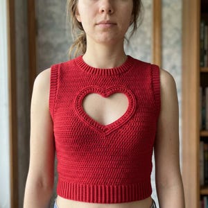 May include: A bright red sleeveless crochet crop top featuring a prominent heart-shaped cutout on the chest. The garment is constructed with a dense stitch pattern, a ribbed crew neckline, ribbed armholes, and a wide ribbed hem at the waist.