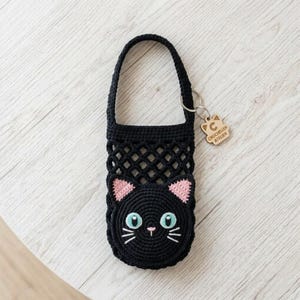 May include: A black crocheted bottle holder featuring a cat face design with light blue eyes, pink inner ears, and white whiskers. The holder has a top handle and a small wooden tag attached to the side that reads CROCHELLER ATELIER.
