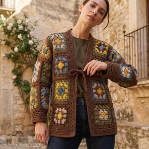 May include: A brown crochet cardigan constructed from multiple square motifs featuring yellow, blue, and green floral patterns. The garment has long sleeves, a V-neckline, and a front tie closure made of matching brown yarn, worn over a dark top.