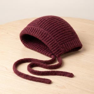 May include: A deep burgundy knit bonnet featuring a ribbed texture throughout the fabric. The headwear includes a rounded crown and a wide, folded brim that frames the face, with two long, matching knitted ties extending from the base for fastening.
