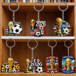 Könnte beinhalten: Eine Sammlung von sieben Metallschlüsselanhängern zum Thema Fußball-Weltmeisterschaft 2026. Jeder Schlüsselanhänger zeigt einen goldenen Pokal, einen schwarz-weißen Fußball und das Jahr 2026. Verschiedene Nationalflaggen und Wahrzeichen sind integriert.