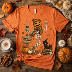 May include: An orange short-sleeve t-shirt featuring a graphic of a skeleton with a pumpkin head wearing a scarf and beanie, holding a coffee cup. The design includes a signpost reading Sweater Weather, Pumpkin Patch, Happy Haunts, and a black cat.