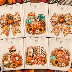 May include: Six cream-colored t-shirts featuring autumn-themed graphic prints. Designs include pumpkins with large bows, sunflowers, a duck wearing sunglasses and a scarf, and the word FALL in patterned letters surrounded by seasonal foliage.