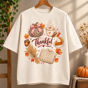 May include: A cream-colored t-shirt featuring a central graphic with the word Thankful in cursive script. The design includes a leopard print pumpkin with a pink bow, a mug of coffee, a slice of pumpkin pie, an acorn, wheat stalks, and a sweater.