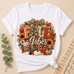 May include: A white short-sleeved t-shirt featuring a graphic print with the text FALL VIBES. The design includes sunflowers, autumn leaves, a plaid bow, pumpkins, and a coffee cup with a heart detail, arranged in a circular seasonal motif.