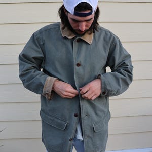 May include: A sage green canvas jacket featuring a contrasting tan corduroy collar and matching tan corduroy sleeve cuffs. The jacket includes a front button closure with dark brown buttons and two large patch pockets at the waist area.