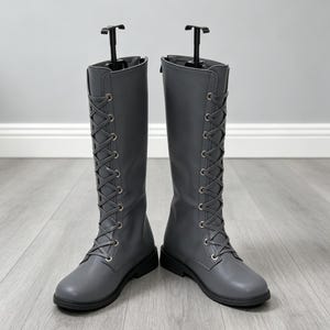 May include: A pair of knee-high grey boots featuring a front lace-up design with silver-toned metal eyelets. The boots have a smooth, matte finish, a rounded toe, and a low heel. Each boot is supported by a black plastic vertical shoe tree insert.
