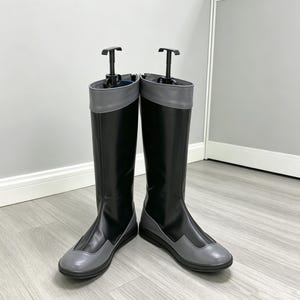 以下が含まれることがあります： 黒いシャフトにグレーのつま先、かかと、トップカフを組み合わせた膝丈のブーツ一足。ブーツは滑らかな質感で、形状を維持するために黒いプラスチック製のブーツキーパーが挿入されており、直立した状態を保っています。