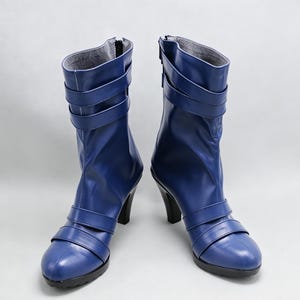Könnte beinhalten: Ein Paar wadenhohe Stiefel in leuchtendem Blau mit glatter Oberfläche, abgerundeter Zehenpartie und hohem Absatz. Jeder Stiefel ist mit zwei horizontalen Riemen über dem Knöchel und zwei Riemen am Schaft sowie einem schwarzen Reißverschluss versehen.