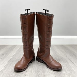 Pode incluir: Um par de botas castanhas até ao joelho com acabamento liso, biqueira arredondada e uma fila vertical de cinco tiras horizontais decorativas na parte frontal do cano. As botas são suportadas por formas de plástico preto.