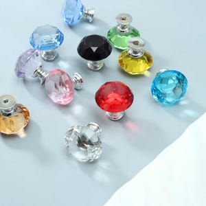 Puede incluir: Una colección de pomos para armarios con facetas en forma de diamante en varios colores, incluyendo rojo, azul, amarillo, verde, negro, rosa, morado, naranja y transparente. Cada pomo tiene una base de metal plateado y una parte superior de cristal.