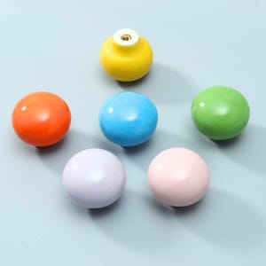 May include: A collection of six round, smooth cabinet knobs in a variety of solid colors including yellow, orange, blue, green, lavender, and light pink. Each knob features a glossy finish and a central metal threaded hole for mounting hardware.