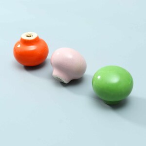 May include: Three round cabinet knobs in glossy orange, matte pastel pink, and glossy lime green. Each knob features a smooth, rounded surface and a central metal threaded insert on the underside for mounting, showcasing a simple, functional design.