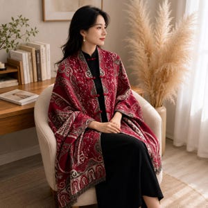 May include: A deep red shawl featuring an intricate white and grey paisley pattern. The garment includes a high collar and is secured at the front with three traditional red fabric toggle closures. The edges are finished with a subtle fringe detail.