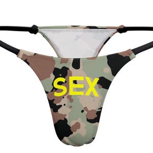 Op de afbeelding: Een string met een camouflagepatroon in tinten groen, bruin, zwart en beige. Het woord SEX is in felgele hoofdletters op de voorkant gedrukt. Het kledingstuk is voorzien van dunne zwarte elastische zijbandjes en een zwarte afwerking.