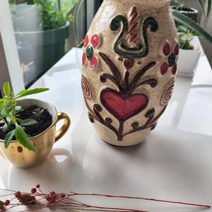 Op de afbeelding: Een keramieken vaas met een crèmekleurig gespikkeld glazuur, voorzien van reliëfmotieven van een centraal rood hart, donkerbruine krullende ranken en rode en groene bloemen. Daarnaast staat een klein goudkleurig kopje met een groene plant.