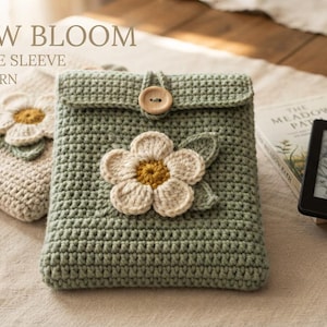 Op de afbeelding: Een saliegroene gehaakte hoes voor boeken en Kindle met een grote witte bloemapplicatie met een geel hart, een houten knoopsluiting en een bijpassende beige hoes op de achtergrond. De tekst luidt MEADOW BLOOM BOOK & KINDLE SLEEVE CROCHET PATTERN.