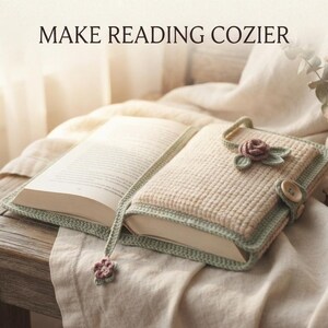 Op de afbeelding: Een beige gehaakte boekomslag met een lichtgroene rand, voorzien van een kleine gehaakte roos en bladversiering, een houten knoopsluiting en een bijpassende bladwijzer met een bloembedel. De tekst MAKE READING COZIER is zichtbaar.