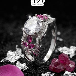 Op de afbeelding: Een zilverkleurige ring met een grote, ronde, heldere middensteen, omringd door kleinere, dieproze markiesgeslepen stenen en kleine heldere accentstenen. De ringband heeft een ingewikkeld, opengewerkt ontwerp. Het logo DB is bovenaan zichtbaar.