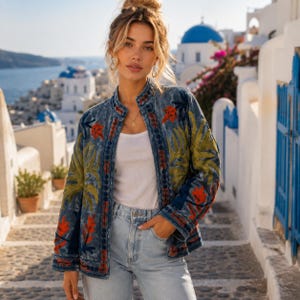 May include: A blue denim jacket featuring a mandarin collar and long sleeves, adorned with vibrant embroidered patterns in shades of olive green and orange. The jacket is worn open over a plain white top and light blue denim jeans.