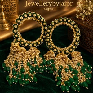 May include: Two gold-tone bangles featuring a circular design with dark green enamel accents and small white floral motifs. Each bangle is adorned with multiple dangling gold chains, small white beads, and dark green cylindrical beads. Text: Jewellerybyjaipr.