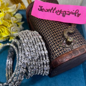 Könnte beinhalten: Ein Set aus silberfarbenen Armreifen mit abwechselnd angeordneten Baguette- und runden klaren Steinen. Ein rosa Zettel mit der Aufschrift Jewellerybyjaily liegt auf einer dunkelbraunen, strukturierten Schatulle mit Metallverschluss.