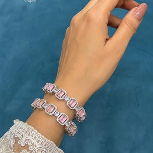 May include: Two silver-toned bracelets featuring rectangular pink stones and small clear accent stones. The text JEWELLERYBYJAIPR is visible in the upper left corner. The bracelets are worn on a wrist with a white lace sleeve cuff visible.