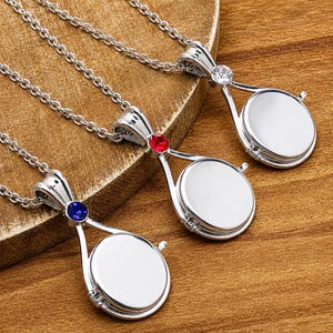 Può includere: Tre collane con medaglione in metallo color argento, ciascuna con un ciondolo circolare dalla superficie liscia e vuota. La parte superiore di ogni ciondolo è decorata con una singola pietra sfaccettata blu, rossa o trasparente su una catena.