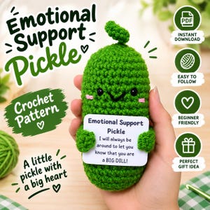 Op de afbeelding: Een groene gehaakte augurk met een lachend gezicht, roze wangen en kleine armen die een wit bordje vasthoudt met de tekst: Emotional Support Pickle, I will always be around to let you know that you are a BIG DILL! De tekst Emotional Support Pickle Crochet Pattern is zichtbaar.