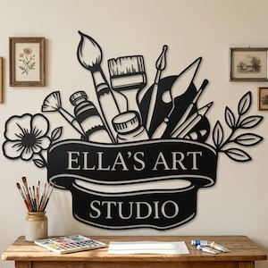 May include: A black metal wall sign featuring the text ELLA'S ART STUDIO on a banner, surrounded by silhouettes of paintbrushes, a palette, a flower, and leaves. The sign is mounted on a wall above a wooden desk with art supplies.