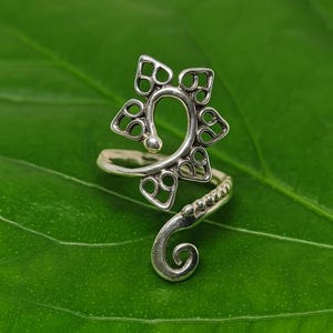 Op de afbeelding: Een zilveren ring met een centraal spiraalontwerp, geaccentueerd door een reeks kleine hartvormige uitsparingen in een cirkelvormig patroon. De band van de ring buigt in een tweede spiraal, versierd met een rij kleine verhoogde zilveren bolletjes.