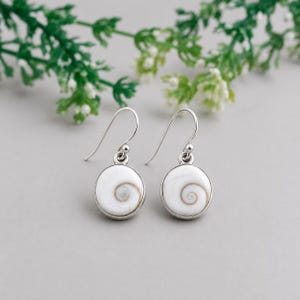May include: A pair of dangle earrings featuring circular white shiva shell inlays set in polished silver-toned metal. Each earring has a curved hook wire and a round pendant displaying a natural spiral pattern in shades of white and light brown.
