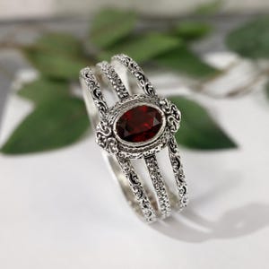 May include: A silver cuff bracelet featuring three bands with intricate scrollwork and filigree detailing. The center of the bracelet is adorned with a large, oval-shaped, deep red garnet gemstone set within a decorative, textured silver frame.