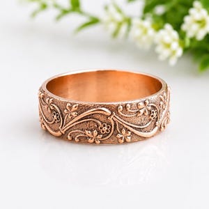 May include: A copper-toned metal ring featuring a textured surface with an embossed floral pattern. The design consists of raised, winding vines and small five-petaled flowers that wrap around the entire circumference of the band.